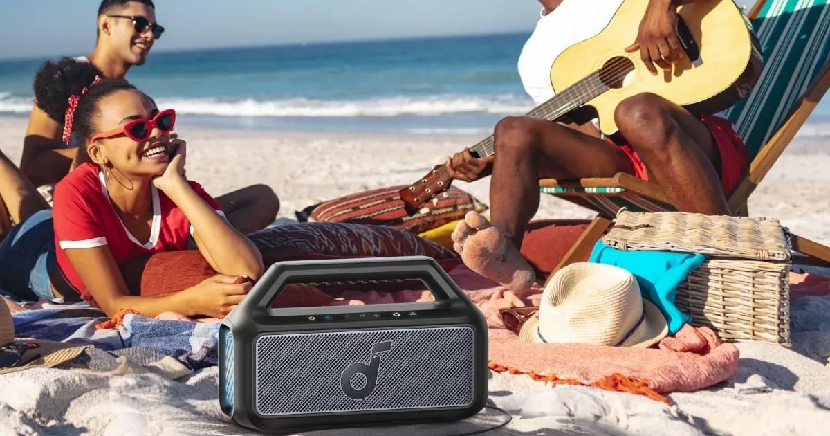 Grupa znajomych na plaży z gitarą, przed nimi głośnik Soundcore Boom 2 na kocu