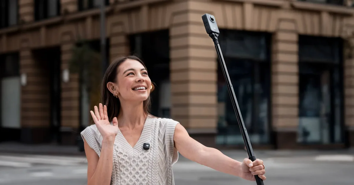 Uśmiechnięta kobieta macha do kamery 360 DJI Osmo 360 zamocowanej na długim kijku na ulicy miasta